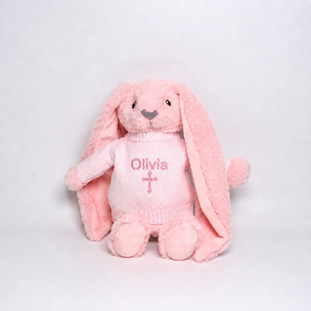 A pink plush bunny toy wearing a matching pink knitted sweater with the name 'Olivia' and a cross embroidered on the front, photographed against a plain light gray background.