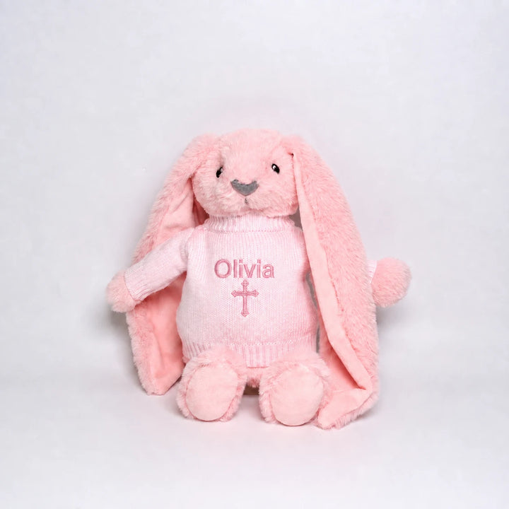 A pink plush bunny toy wearing a matching pink knitted sweater with the name 'Olivia' and a cross embroidered on the front, photographed against a plain light gray background.