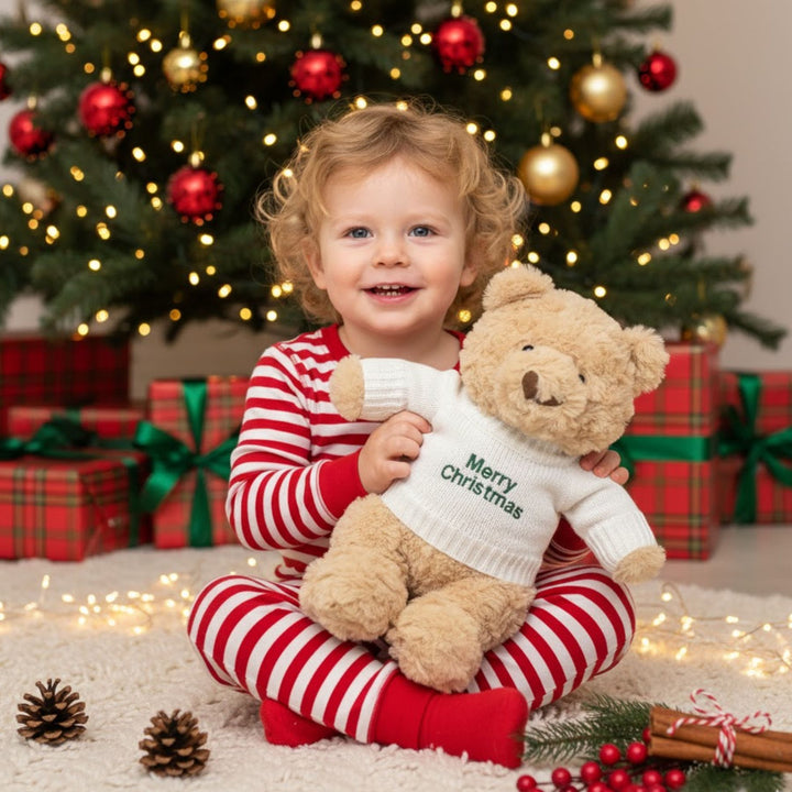 Child in red and white striped pajamas holding a teddy bear with a 'Merry Christmas' sweater in front of a decorated Christmas tree.