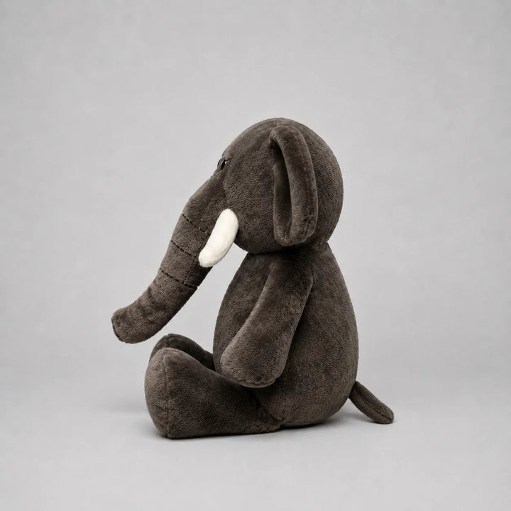 Side view of a grey elephant soft toy with a long trunk, white tusk, floppy ear and oversized feet, photographed on a light grey studio background.