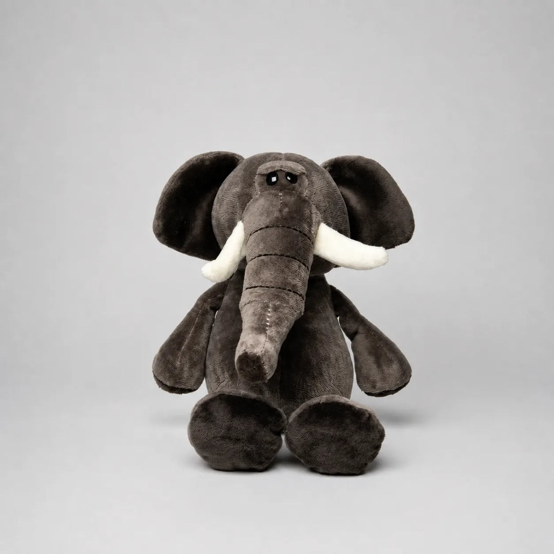 Grey elephant soft toy with a long trunk, large floppy ears, white tusks and oversized feet, photographed from the front on a light grey studio background.