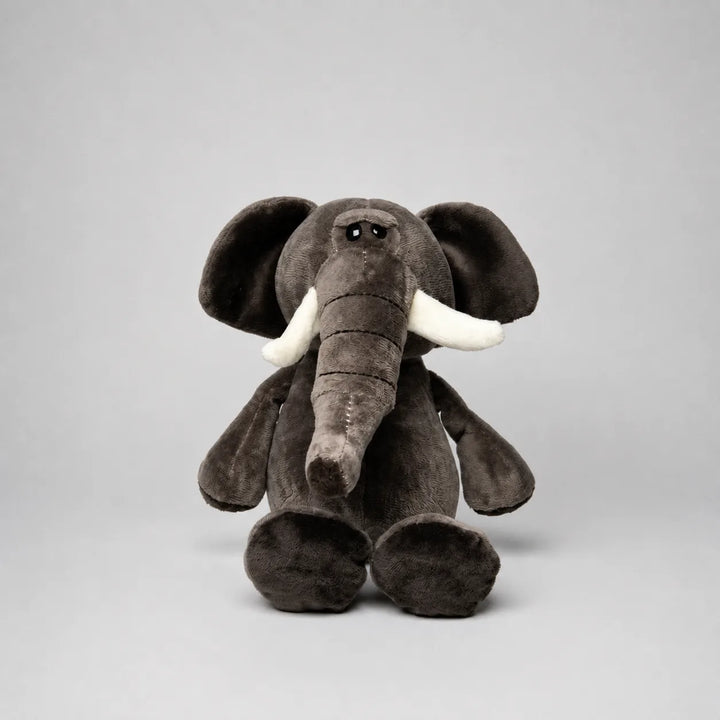 Grey elephant soft toy with a long trunk, large floppy ears, white tusks and oversized feet, photographed from the front on a light grey studio background.
