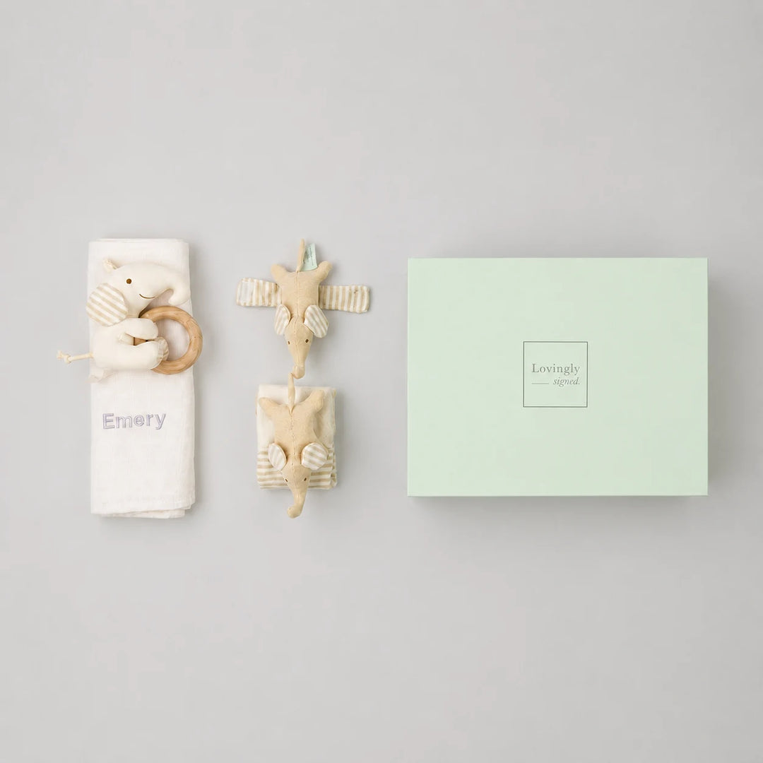 A baby gift set featuring a white bamboo swaddle with a wooden elephant teether and 'Emery' printed on it, two plush animal teething toys, and a mint green gift box.