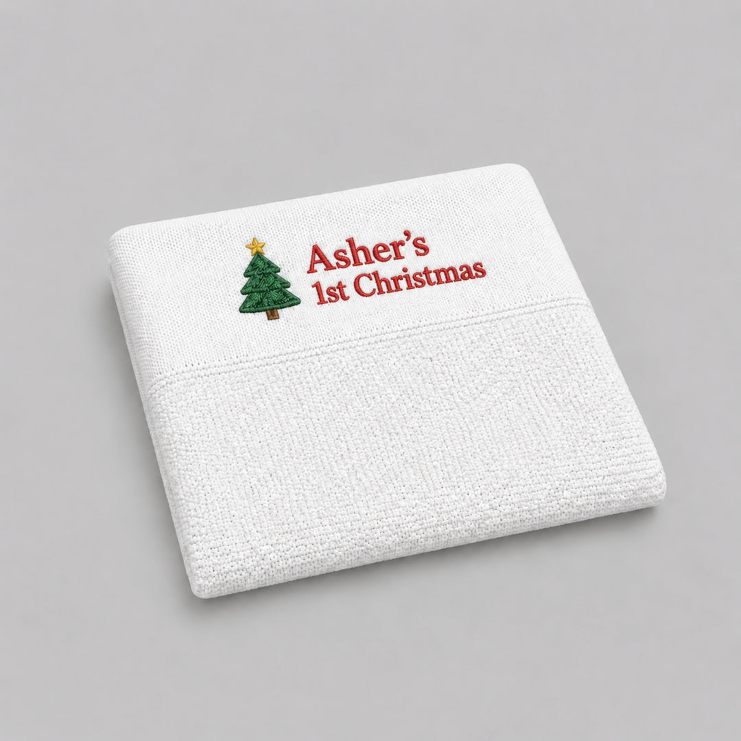 White personalised knitted Christmas baby blanket embroidered with a Christmas tree and “Asher’s 1st Christmas” in red, photographed on a light grey studio background.