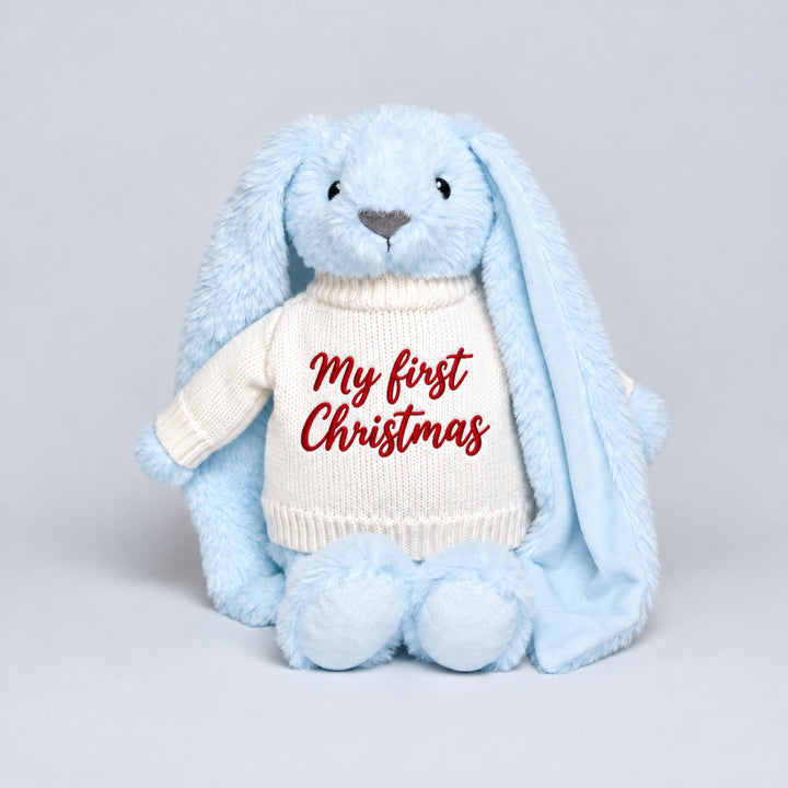 Blue bunny soft toy wearing a cream knitted jumper embroidered with “My First Christmas” in red, photographed on a light grey studio background.