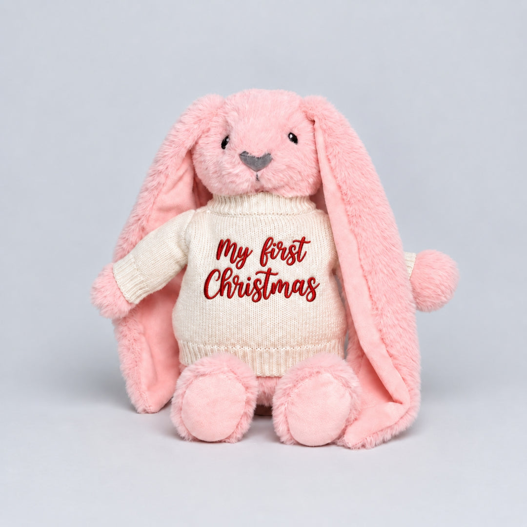 Pink bunny soft toy wearing a cream knitted jumper embroidered with “My First Christmas” in red, photographed on a light grey studio background.