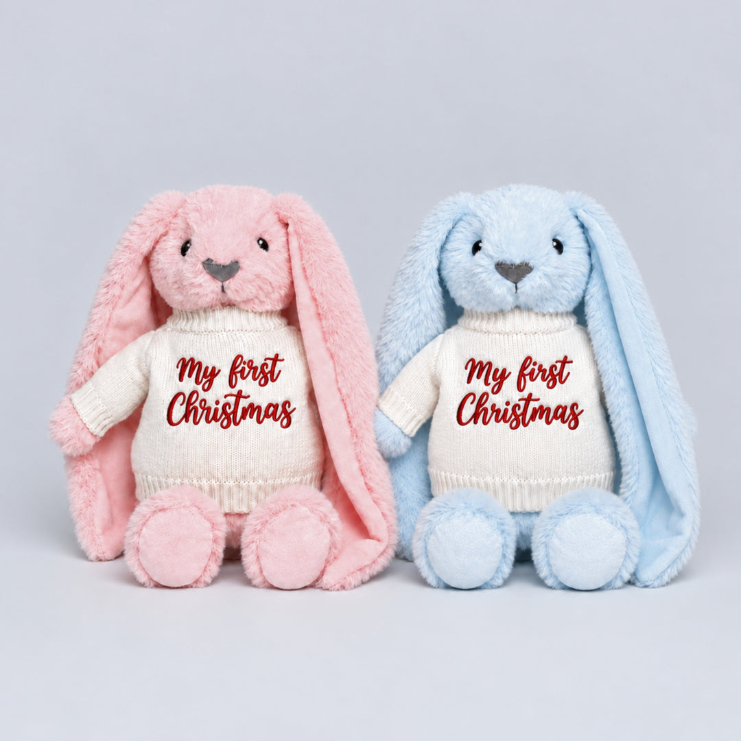 Pink and blue bunny soft toys wearing cream knitted jumpers embroidered with “My First Christmas” in red, photographed together on a light grey studio background.