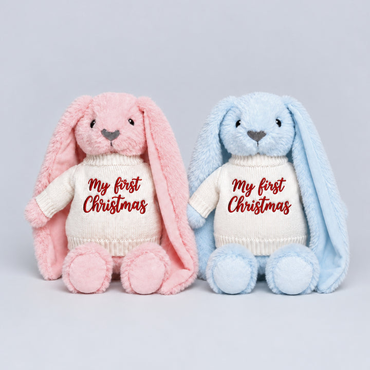 Pink and blue bunny soft toys wearing cream knitted jumpers embroidered with “My First Christmas” in red, photographed together on a light grey studio background.