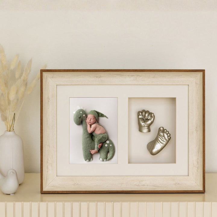 Framed artwork with a baby in a green outfit and dinosaur costume, next to metallic hand and footprints on a neutral background.