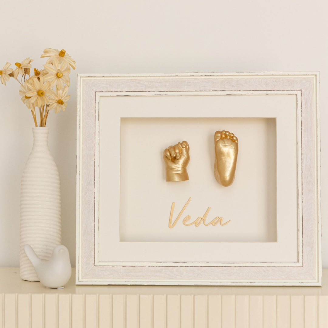 Framed gold hand and footprints with 'Veda' on a white wall with decorative elements.