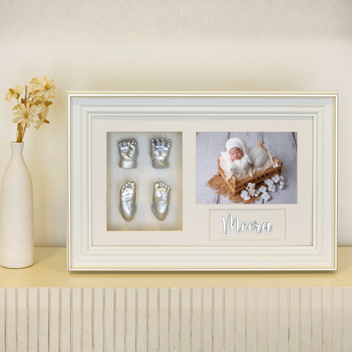 Framed display with baby's feet casts and a photo of a baby named Meera on a light wooden shelf.