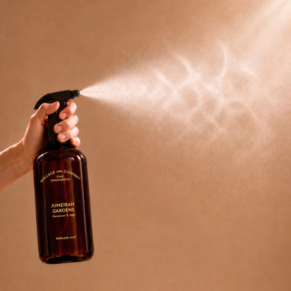 A hand holds a dark amber spray bottle labeled 'Wallace and Cowell Fine Fragrances' and 'Jumeirah Gardens', spraying a fine mist against a warm beige background.