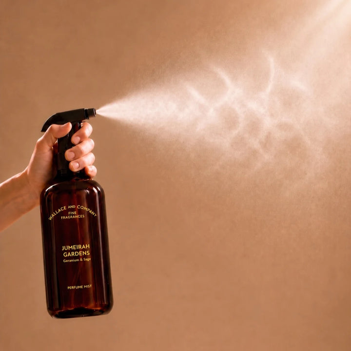 A hand holds a dark amber spray bottle labeled 'Wallace and Cowell Fine Fragrances' and 'Jumeirah Gardens', spraying a fine mist against a warm beige background.