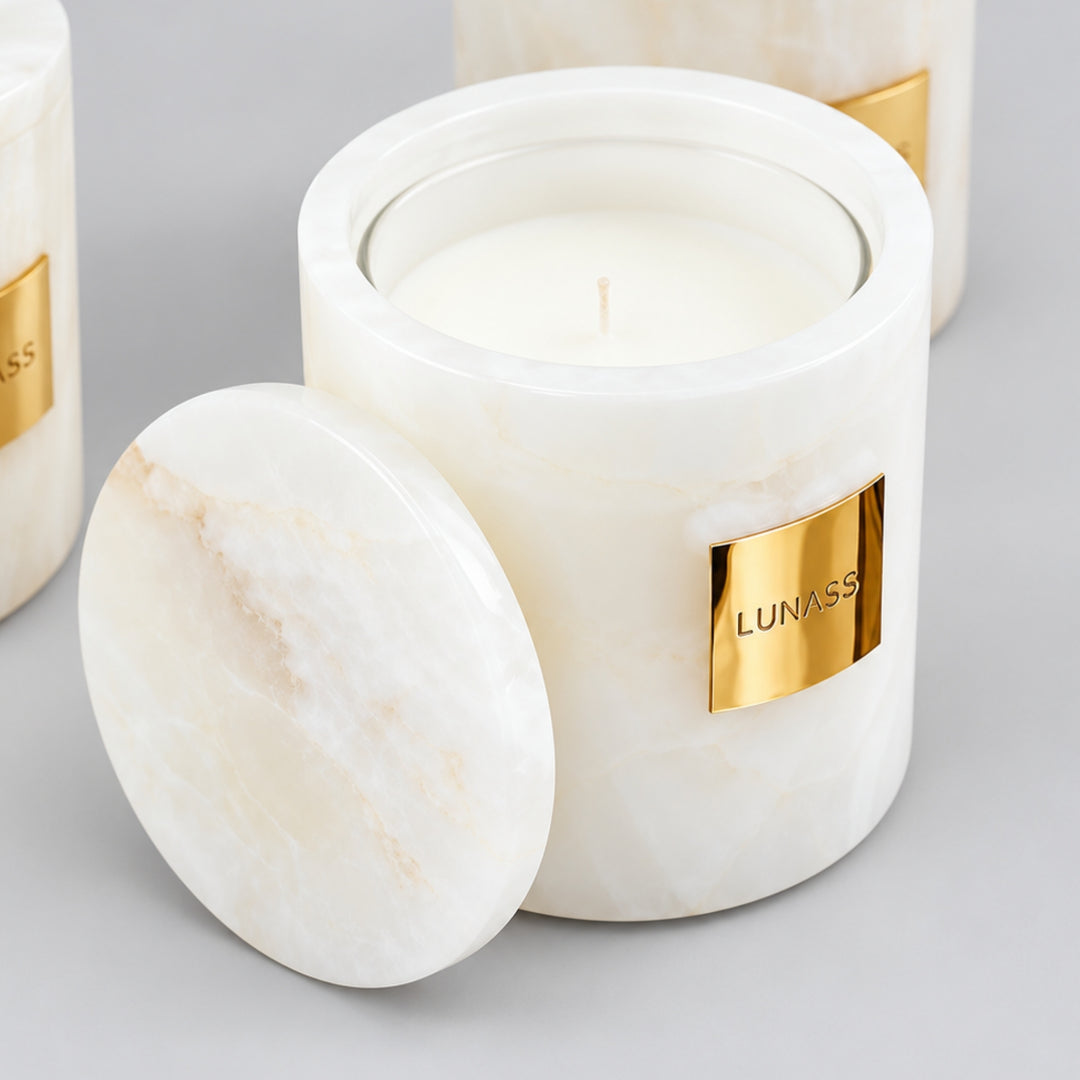 Close-up of a white marble-effect LUNASS candle with a gold label and matching round lid resting in front, with two blurred matching candles in the background on a soft grey backdrop