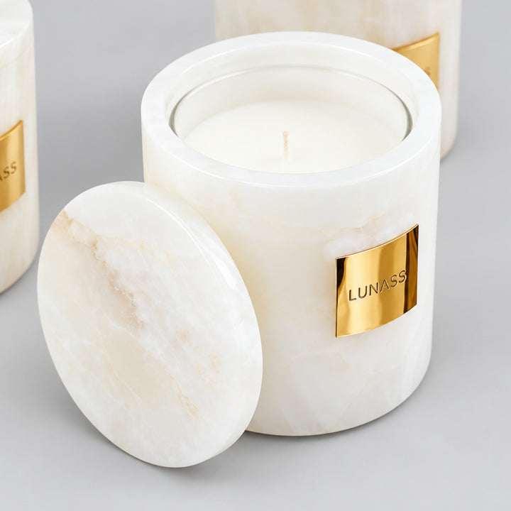 Close-up of a white marble-effect LUNASS candle with a gold label and matching round lid resting in front, with two blurred matching candles in the background on a soft grey backdrop