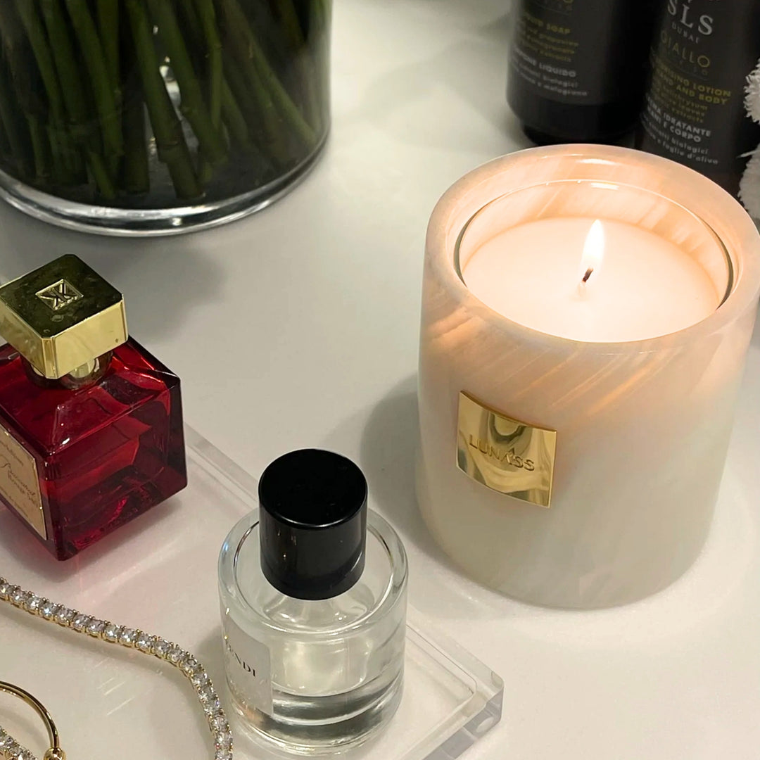 Lit white marble LUNASS candle with a gold label styled on a vanity table beside perfume bottles, jewellery, flowers, and beauty products in a warm, elegant lifestyle setting.