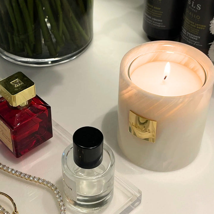 Lit white marble LUNASS candle with a gold label styled on a vanity table beside perfume bottles, jewellery, flowers, and beauty products in a warm, elegant lifestyle setting.