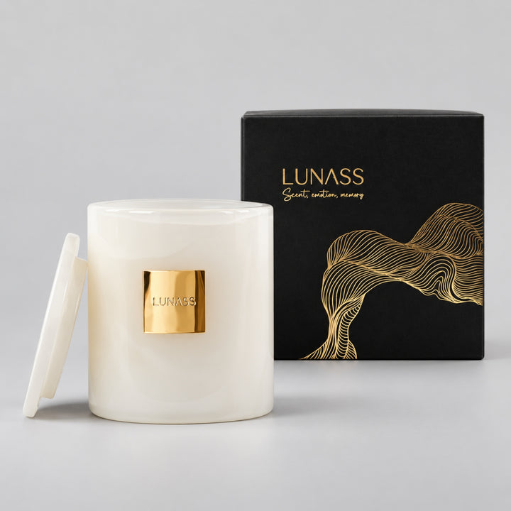White LUNASS luxury candle with a gold label, shown with its lid leaning on the left and a black gift box with gold detailing behind it, photographed on a light grey studio background.