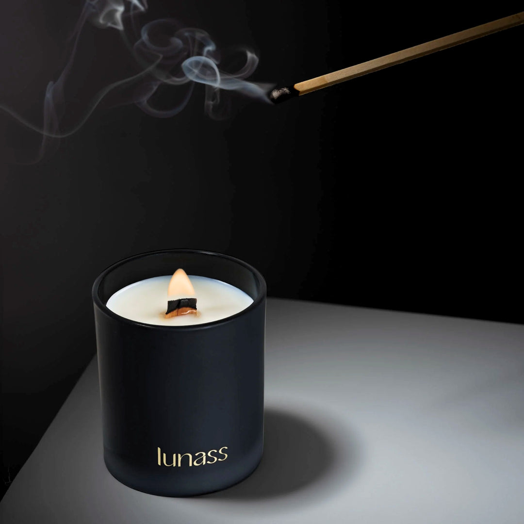Black LUNASS scented candle burning on a dark surface as smoke curls from a freshly extinguished match above.