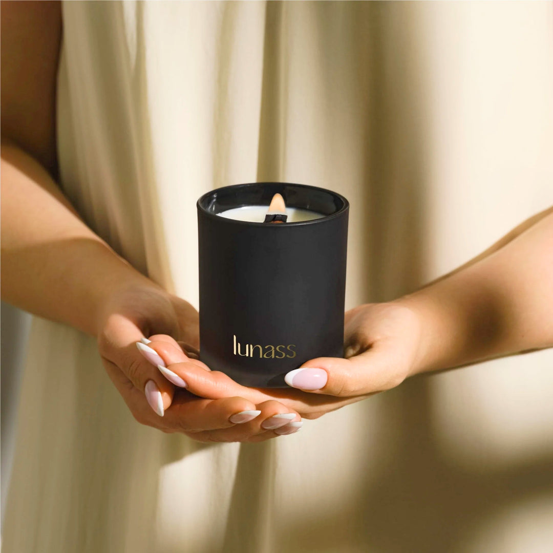 Lifestyle image of a lit black LUNASS scented candle held gently in both hands against a warm neutral background.