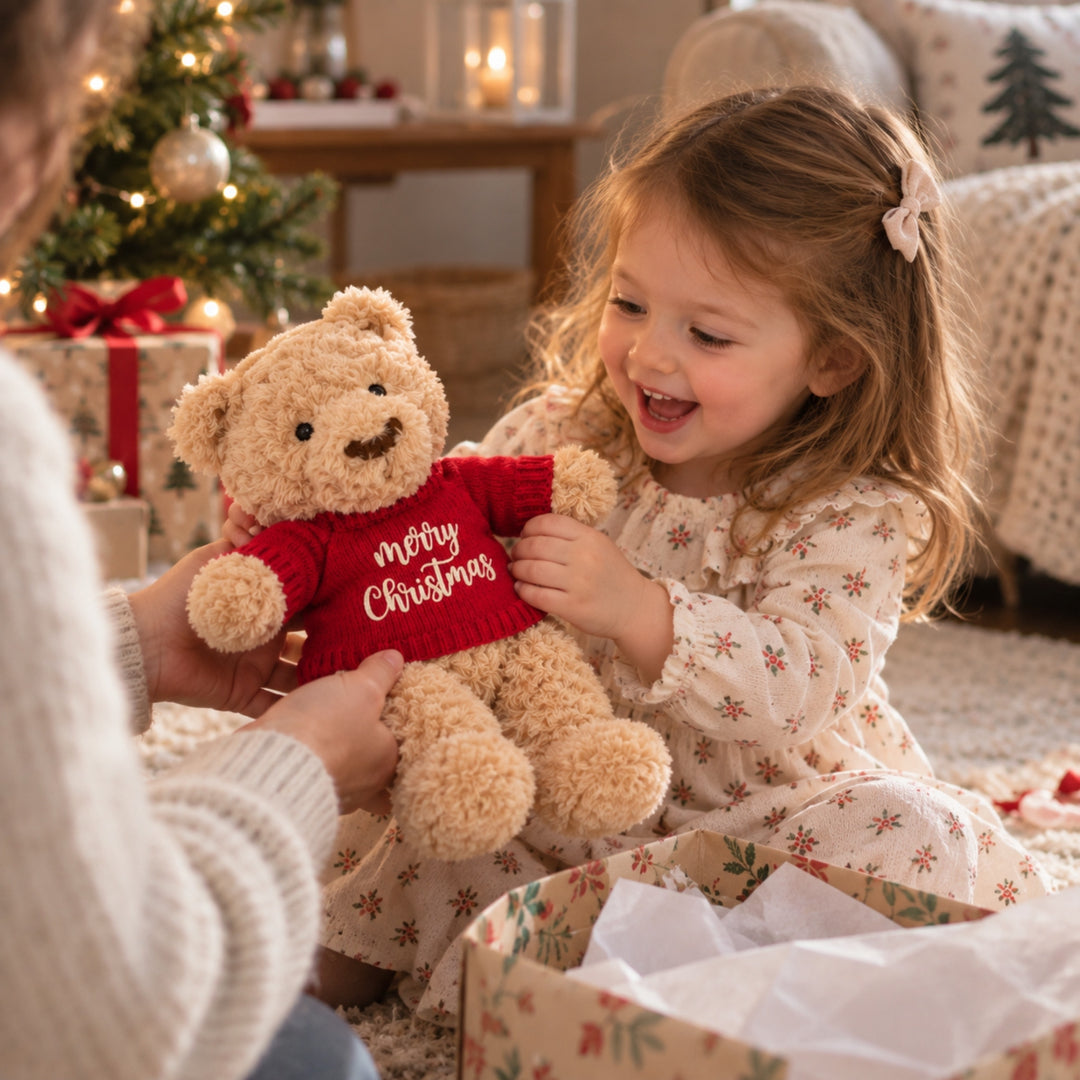 Young girl receiving and cuddling a teddy bear wearing a red knitted “Merry Christmas” jumper beside a decorated Christmas tree and wrapped presents.