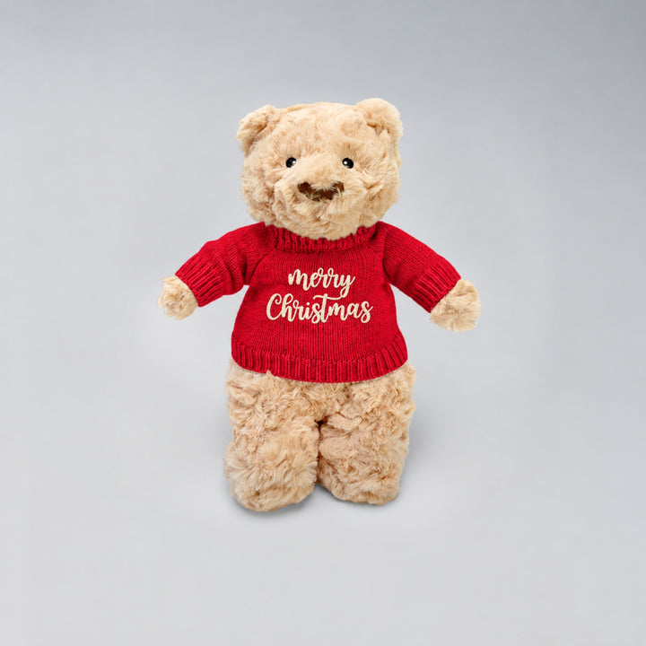 Teddy bear wearing a red knitted jumper embroidered with “Merry Christmas”, photographed on a light grey studio background.