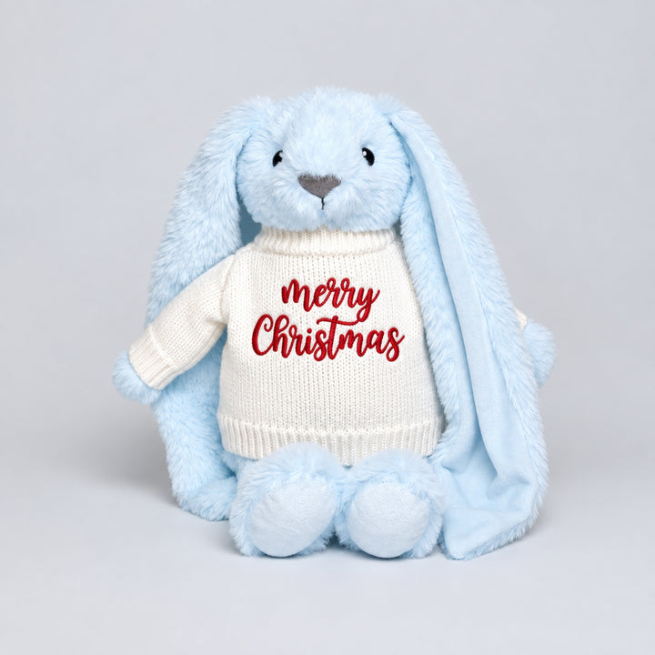 Blue bunny soft toy wearing a cream knitted jumper embroidered with “Merry Christmas” in red, photographed on a light grey studio background.