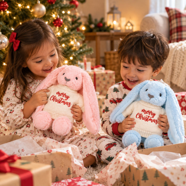 Two young children opening Christmas gifts and cuddling pink and blue bunny soft toys dressed in cream knitted jumpers embroidered with “Merry Christmas”, with a decorated Christmas tree and wrapped presents in the background.