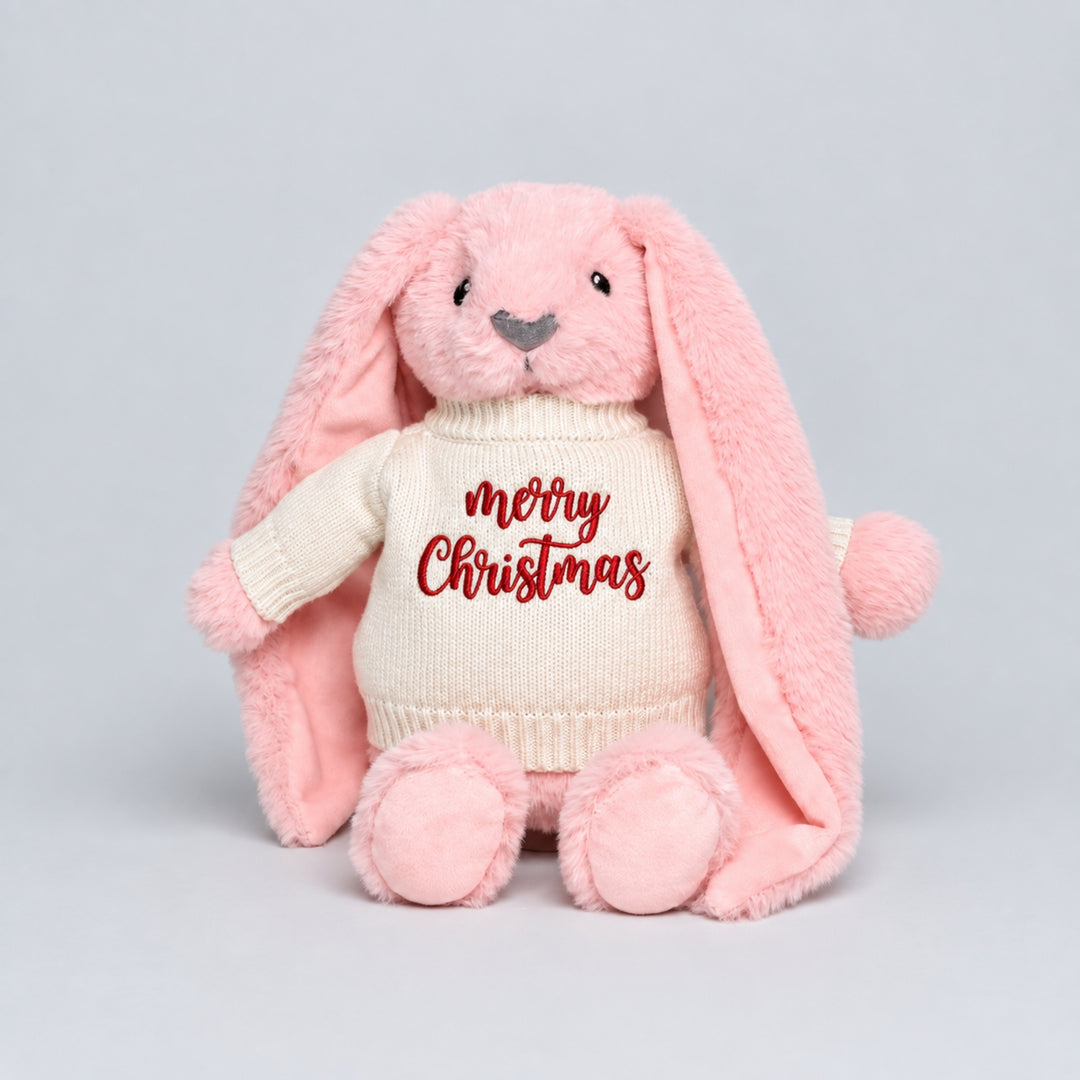 Pink bunny soft toy wearing a cream knitted jumper embroidered with “Merry Christmas” in red, photographed on a light grey studio background.