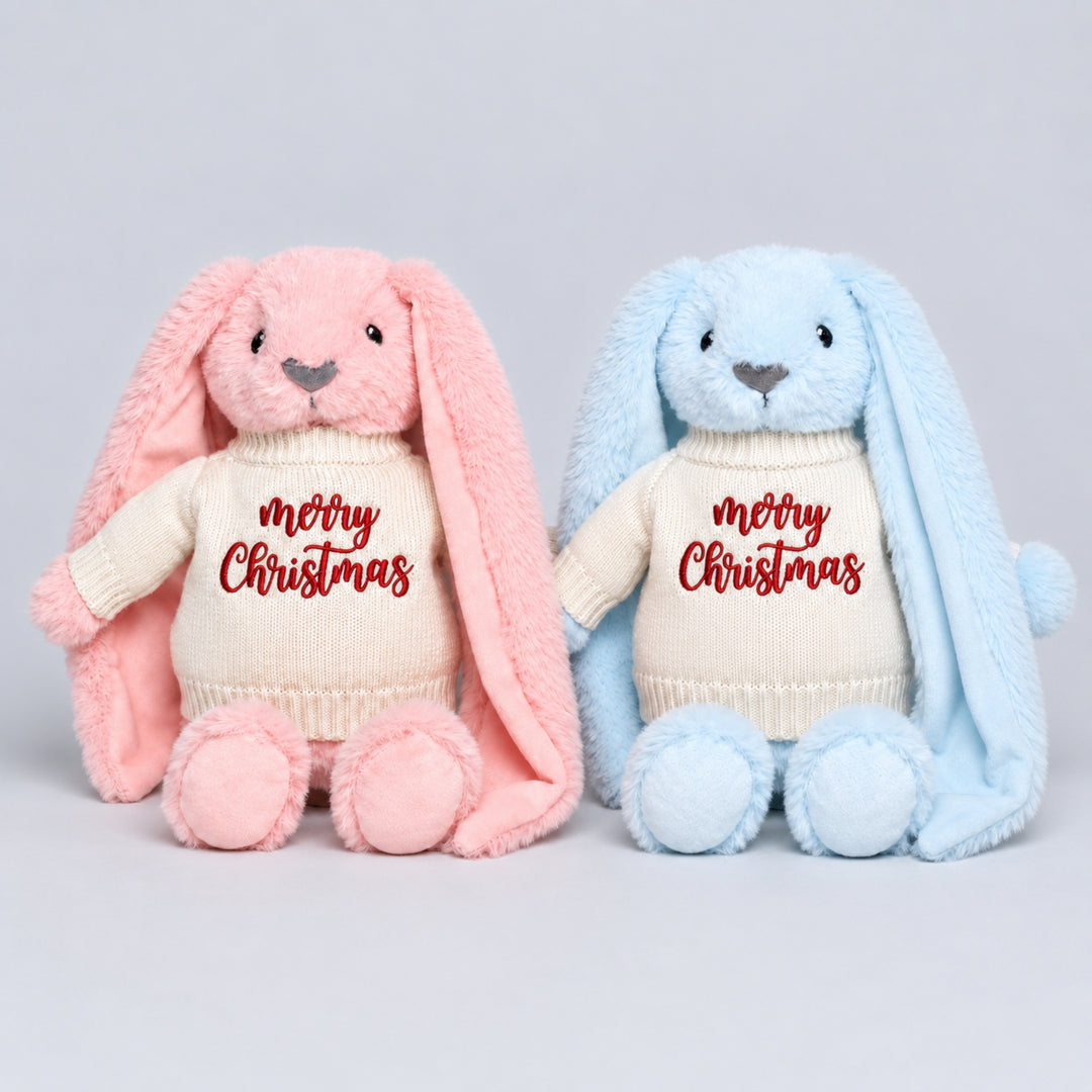 Pink and blue bunny soft toys wearing matching cream knitted “Merry Christmas” jumpers, photographed together on a light grey studio background.
