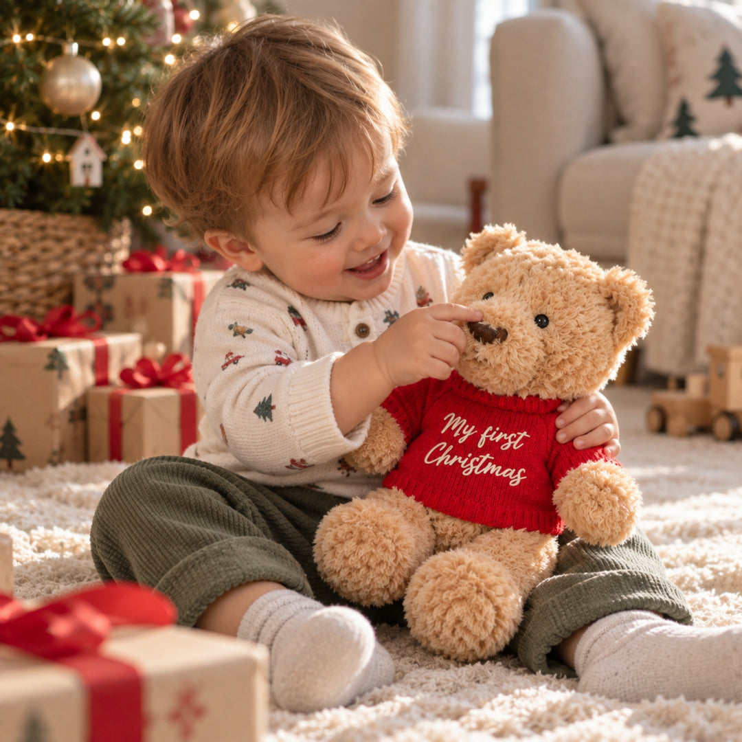 Young child sitting beside a Christmas tree and cuddling a teddy bear wearing a red “My First Christmas” jumper in a cosy festive living room.