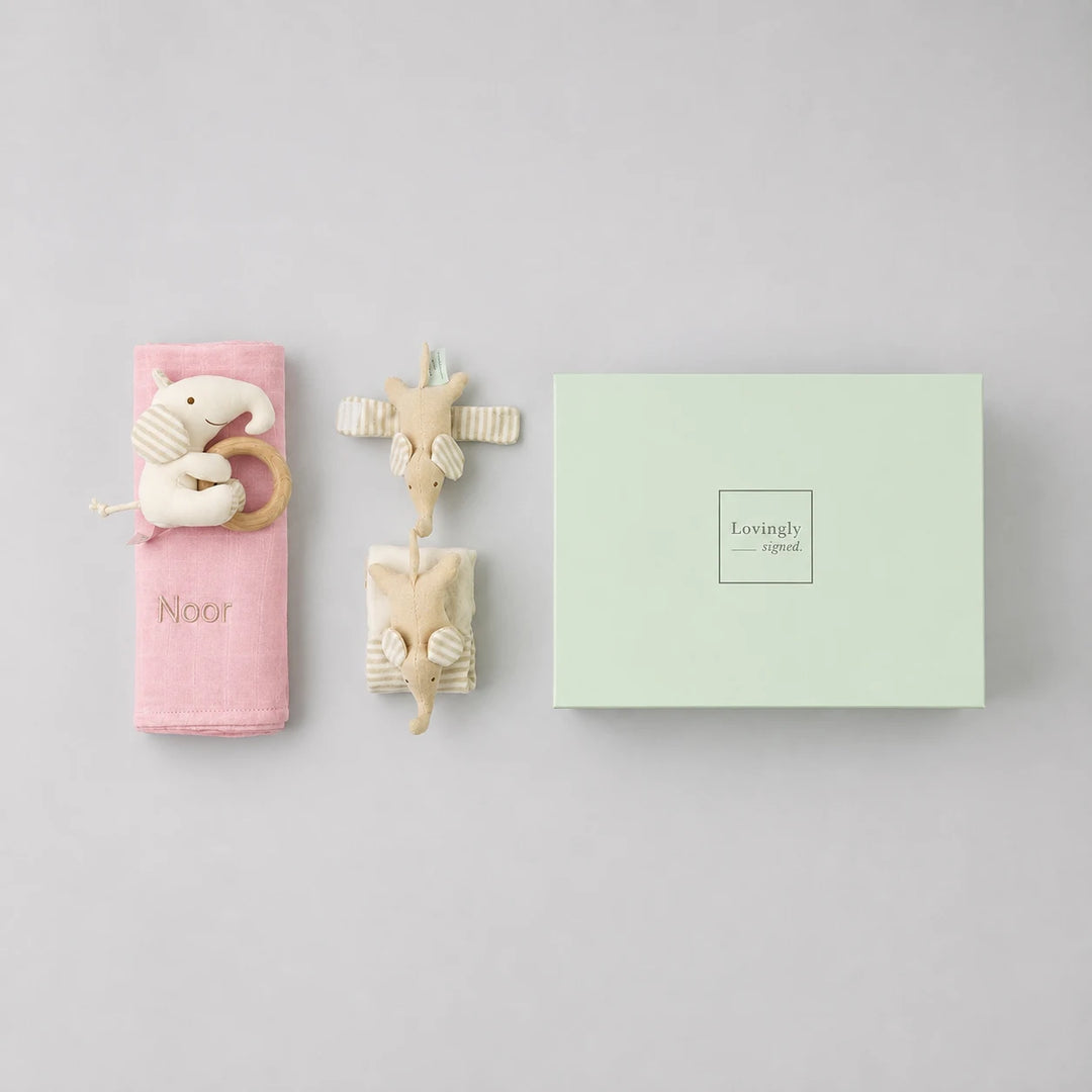 A baby gift set featuring a pink swaddle with a wooden elephant teether and the name 'Emery', and a mint green gift box with 'Lovingly Styled' printed on it, all arranged on a white background.