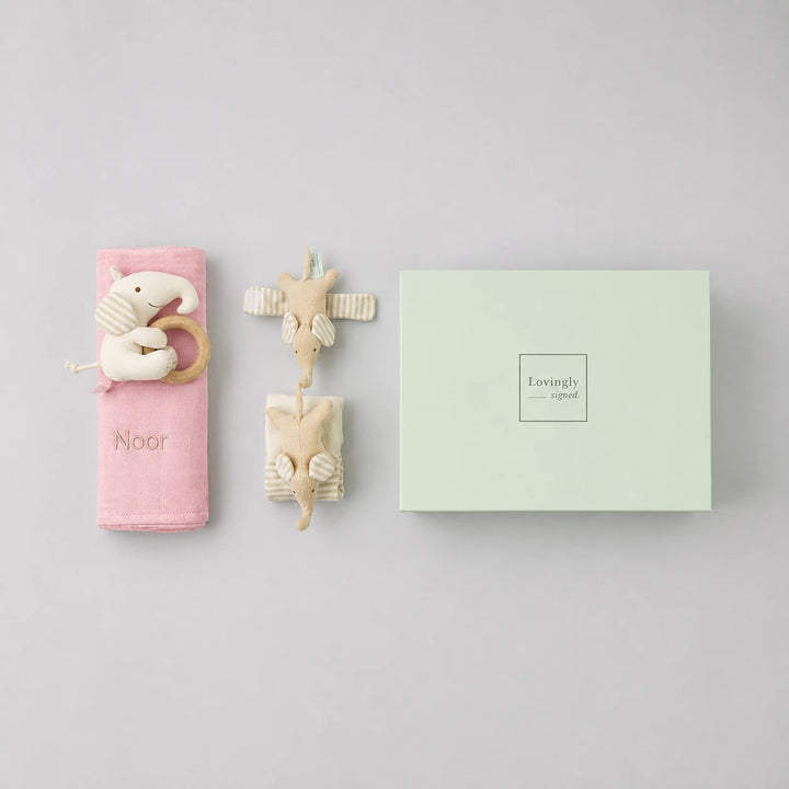 A baby gift set featuring a pink swaddle with a wooden elephant teether and the name 'Emery', and a mint green gift box with 'Lovingly Styled' printed on it, all arranged on a white background.