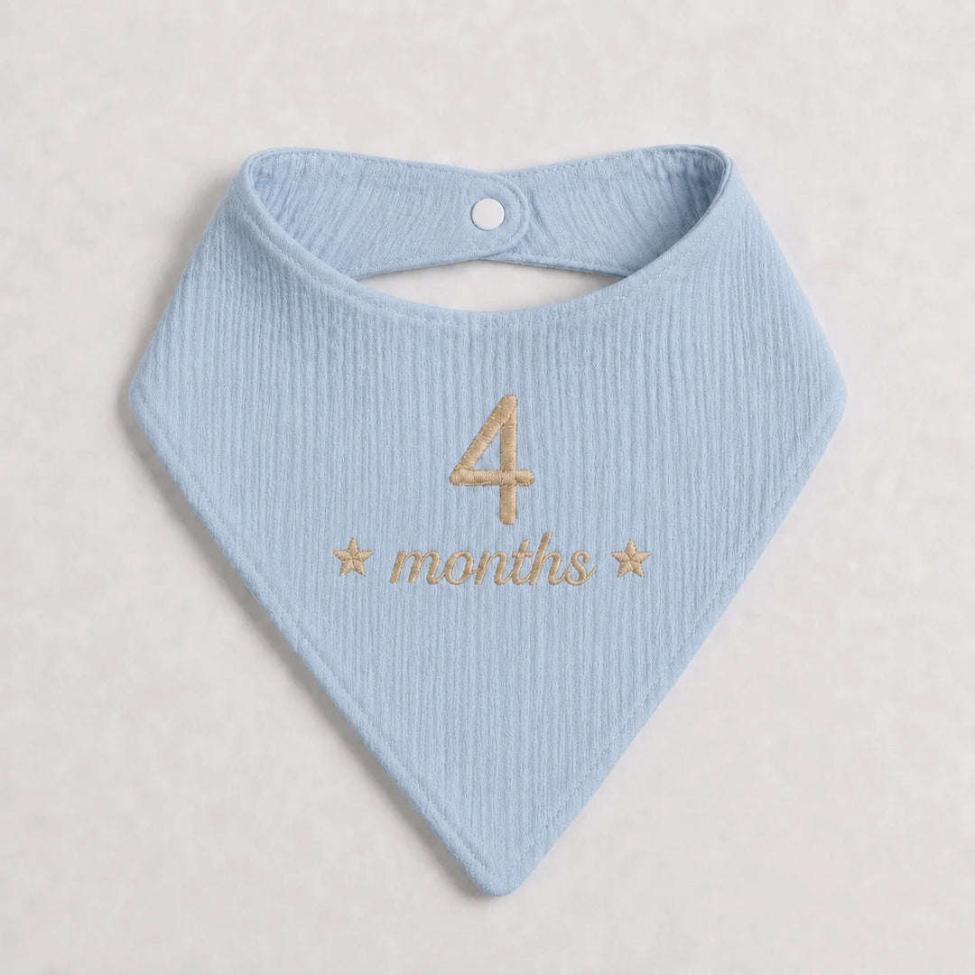 A light blue ribbed cotton bandana bib with gold embroidered text reading '4 months' and small stars, displayed flat on a light beige textured background.