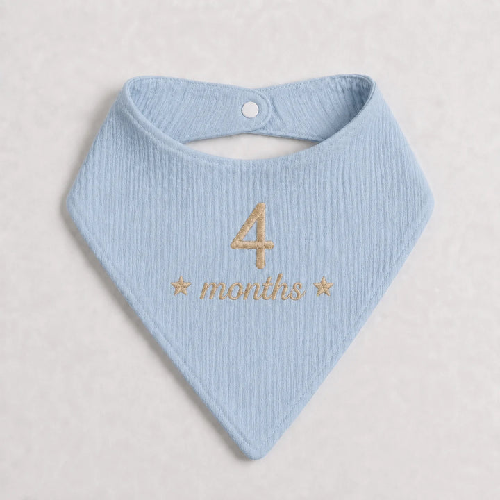 A light blue ribbed cotton bandana bib with gold embroidered text reading '4 months' and small stars, displayed flat on a light beige textured background.