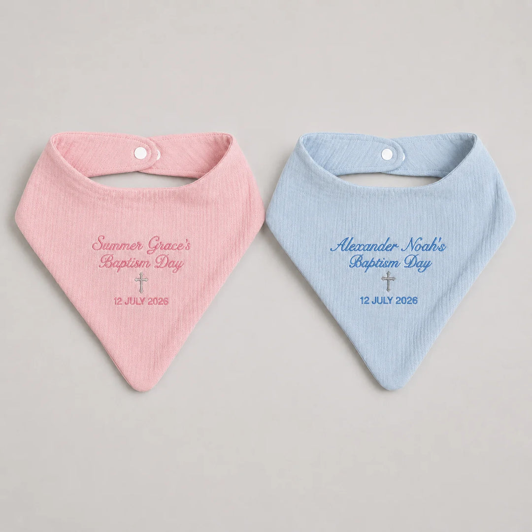 Two personalised baby bandana bibs, one light pink and one light blue, laid flat on a neutral light gray background. Each bib features a snap closure at the top and custom embroidered text and a small cross on the front.