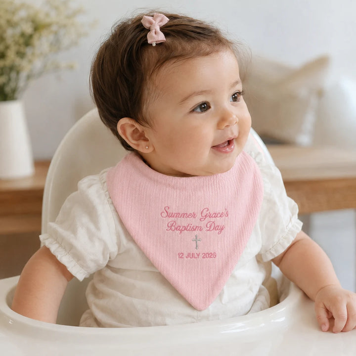 A baby wearing a light pink bib with personalized text 'Summer Grace's Baptism Day 12 JULY 2025' sits in a white high chair. The baby is wearing a white short-sleeve ruffled shirt and has a pink bow in her hair.
