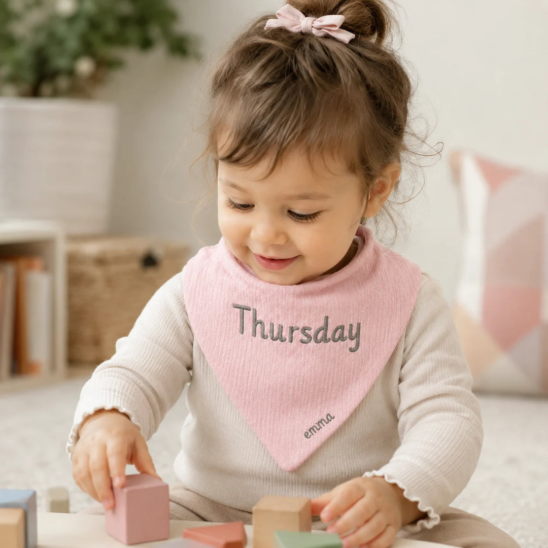 A toddler girl wearing a light pink bandana bib with 'Thursday' and 'emma.' printed on it, sitting on a floor and playing with wooden blocks.