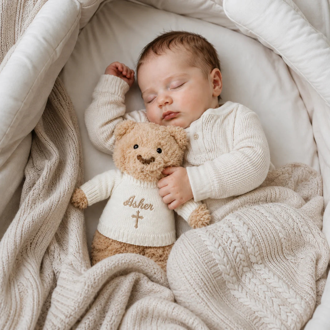 A sleeping newborn baby wearing a cream knit cardigan lies in a soft cream bassinet, hugging a teddy bear wearing a matching cream sweater with the name 'Avery' and a cross embroidered on the front.