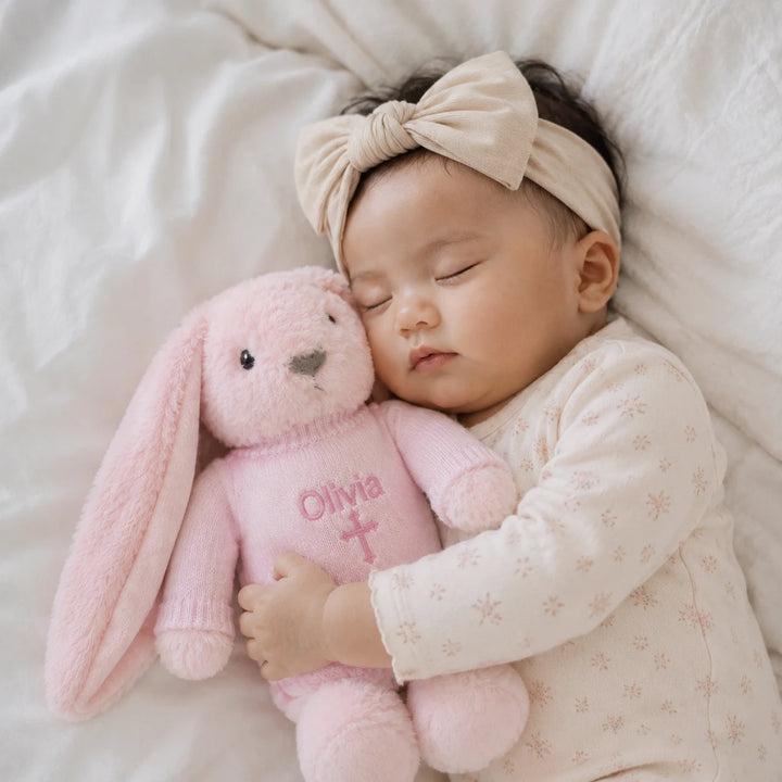 A sleeping newborn baby wearing a cream floral long-sleeve bodysuit and a beige velvet bow headband, cuddling a personalized pink plush bunny toy embroidered with the name 'Olivia' and a cross.