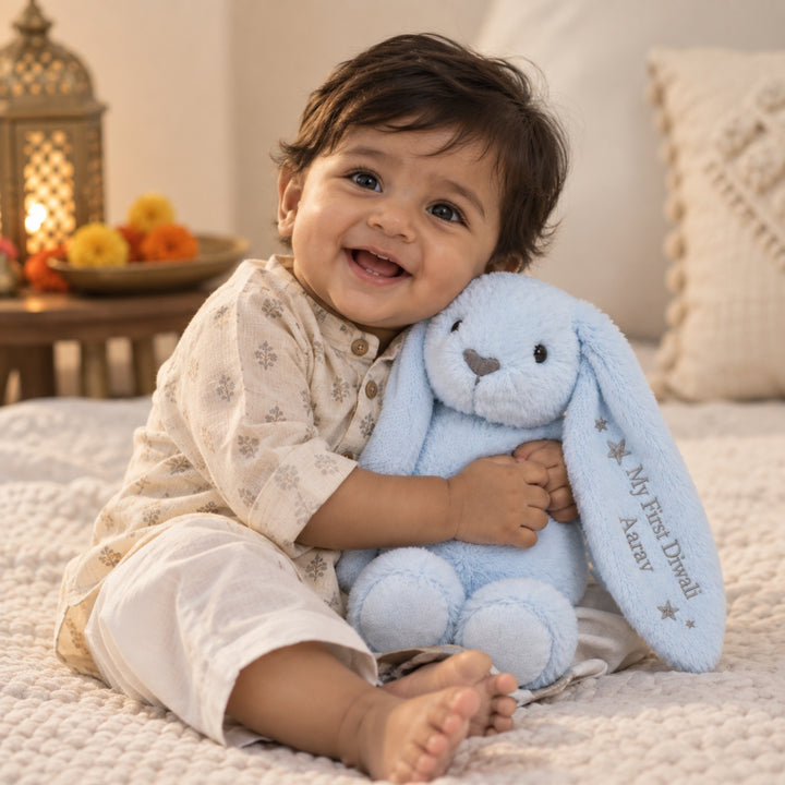 Smiling baby boy cuddling a personalised baby blue “My First Diwali Aarav” bunny soft toy in a warm festive home setting.