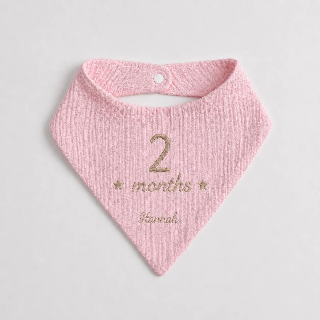 A light pink textured cotton bandana bib with a white snap button, personalized with '2 months Hannah' in beige embroidery on a light gray background.