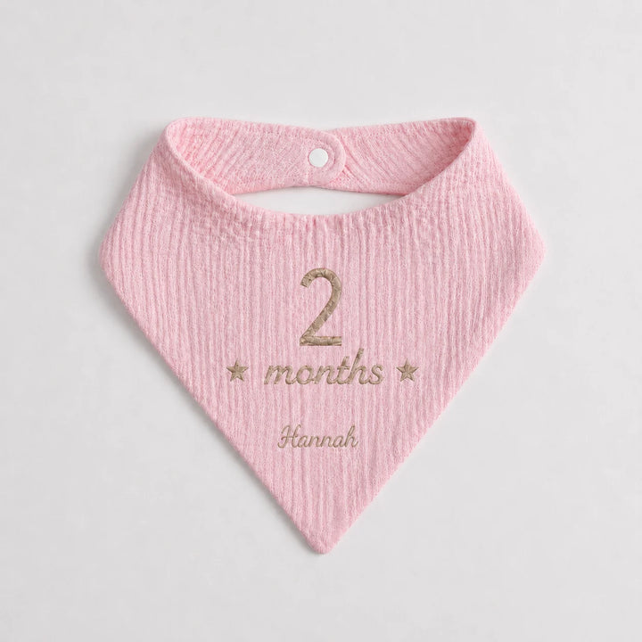 A light pink textured cotton bandana bib with a white snap button, personalized with '2 months Hannah' in beige embroidery on a light gray background.