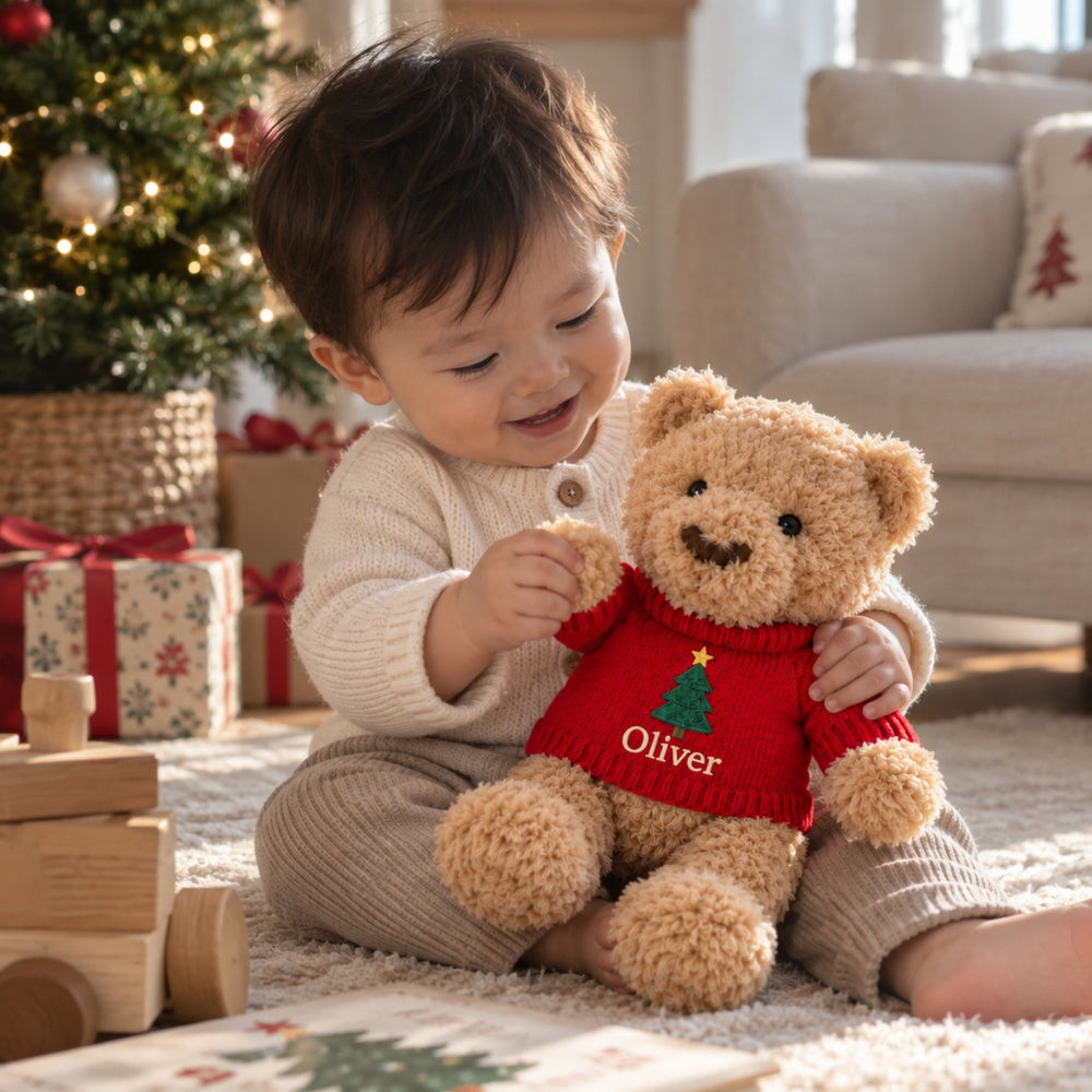Young child in a festive living room cuddling a personalised Christmas teddy bear wearing a red jumper embroidered with a Christmas tree and the name “Oliver”, with a decorated Christmas tree and wrapped presents in the background.