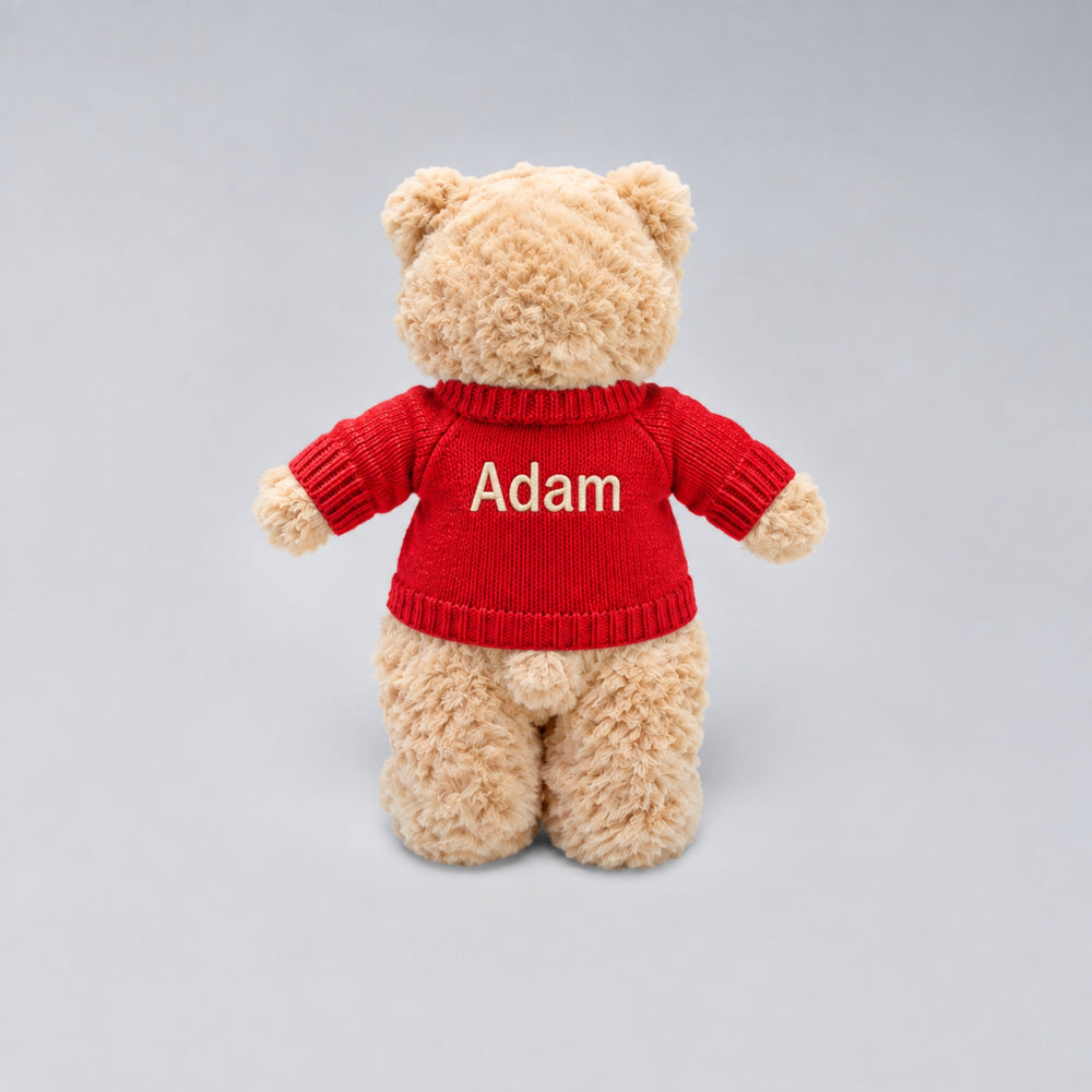 Personalised Christmas teddy bear wearing a red knitted jumper embroidered with the name “Adam” on the back, photographed on a light grey studio background.