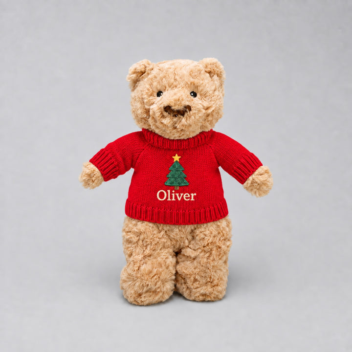 Personalised Christmas teddy bear wearing a red knitted jumper embroidered with a Christmas tree and the name “Oliver”, photographed on a light grey studio background.