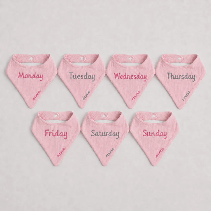 Set of seven pink baby bandannas with days of the week text on a light gray background