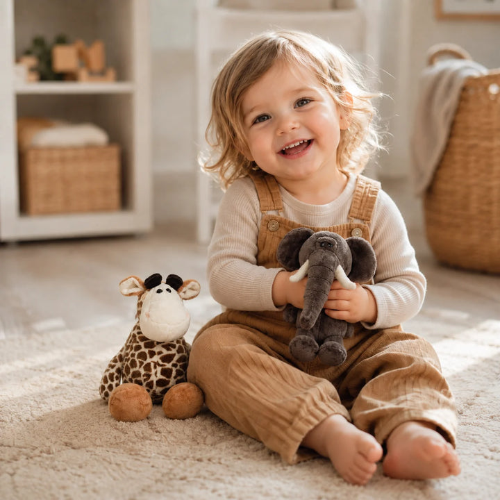 Toddler sitting on a soft rug holding a grey elephant plush toy, with a giraffe soft toy beside them in a warm neutral nursery.