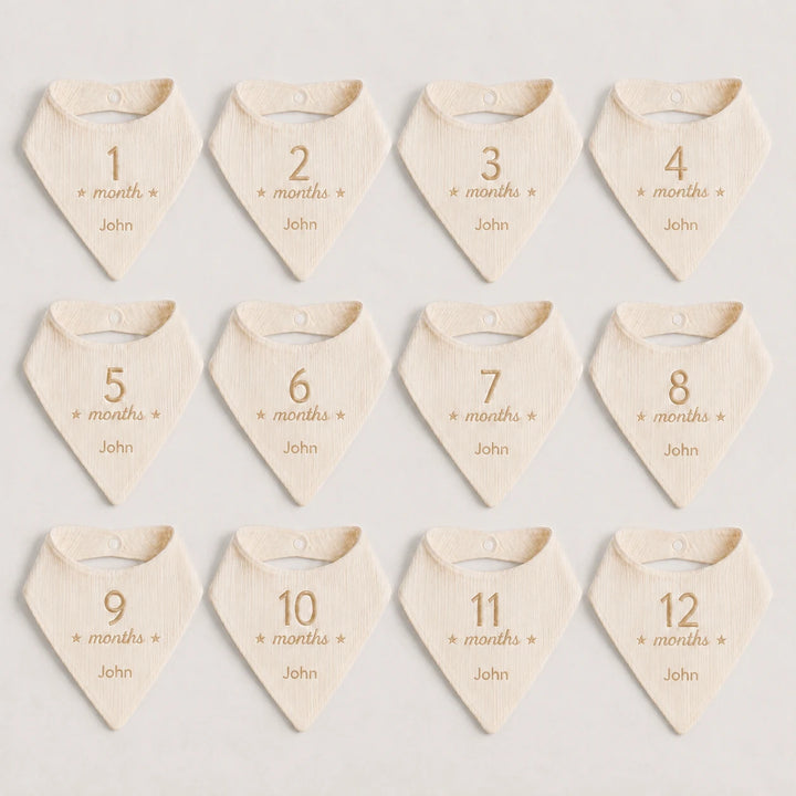 Twelve triangular baby bibs arranged in a 3-by-4 grid on a light neutral background. Each bib is cream-colored with a small gold number from 1 to 12, followed by the word 'months' and small gold stars.