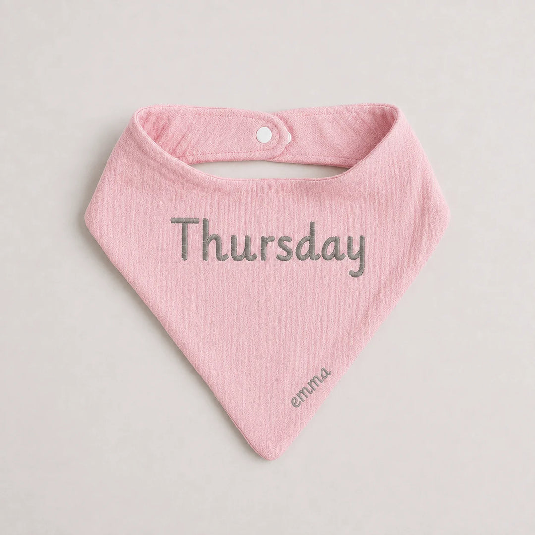 A light pink textured baby bandana bib with 'Thursday' embroidered in gray across the center and 'emma' in a smaller font near the bottom tip, shown on a neutral light gray background.